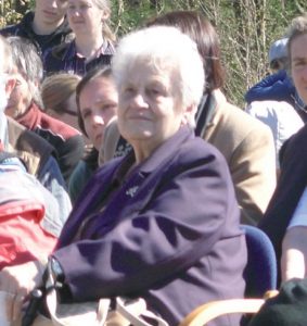 Anni Kupper, eine alte Frau mit weißen kurzen Haaren, sitzt auf einem Stuhl an der frischen Luft. Um sie herum sind weitere Menschen. Sie alle schauen und hören gerade bei etwas zu.