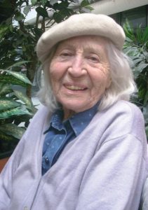 Porträtfoto von Irma Trksak, einer alten lächelnden alten Frau mit schulterlangem grauen Haar und Baskenmütze. Im Hintergrund eine Pflanze.