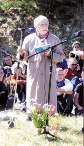 Lucja Barwikowska, eine alte Frau mit konzentriertem Gesichtsausdruck, steht auf einer Wiese und spricht in ein Mikrofon. Vor ihr steht ein Blumenstrauß. Hinter ihr sitzen zuhörende Menschen auf dem Gras.