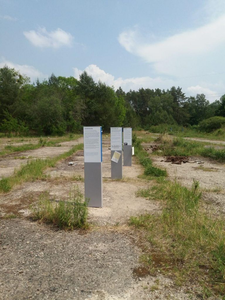 Stelen auf dem Gednekort Uckermark auf Betonplatten auf einer weiten Fläche, mit Bäumen im Hintergrund.
