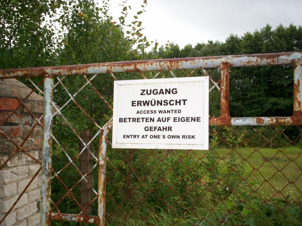 Das Foto zeigt ein Eingangstor zum Gedenkort ehemaliges KZ Uckermark. Auf dem Tot ist ein Schild mit der Aufschrift "Zugang erwünscht/Access wanted. Betreten auf eigene Gefahr/Entry at one´s own risk" zu sehen.